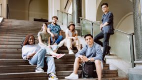 Six international students sitting in a relaxed manner in the hallway of a university. They smile towards the camera.