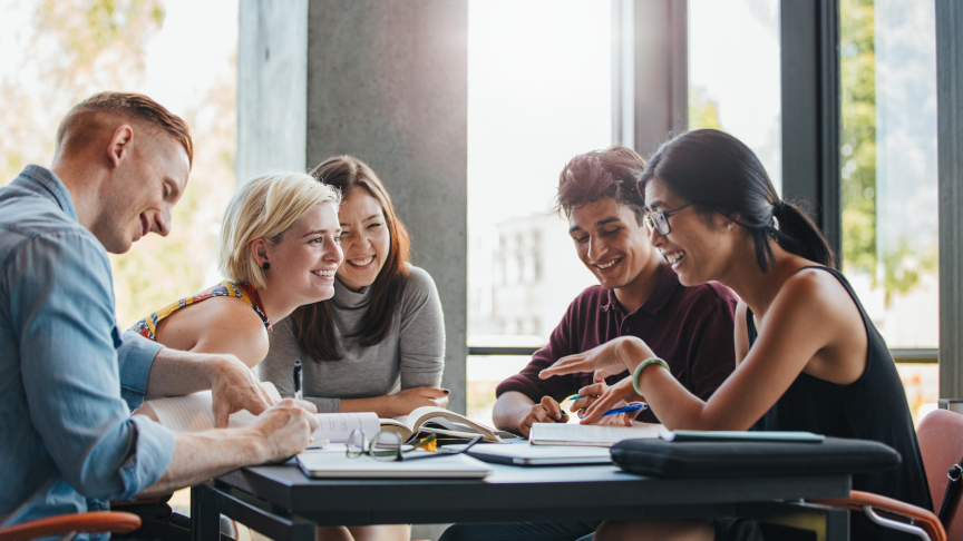 Internationale Studierende sitzen lachend an einem Tisch in der Universität und lernen gemeinsam.