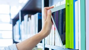 A hand takes a tablet from a bookshelf in the library.